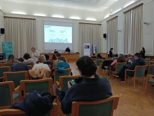 Zbiranje udeležencev pred prvim usposabljanjem. Velika dvorana, v ospredju projektorsko platno in 20+ udeležencev.