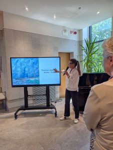 La relatrice descrive un'opera d'arte visualizzata sullo schermo, mentre i partecipanti seguono la presentazione.