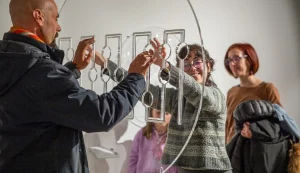 Due visitatori della galleria esplorano con le mani un'installazione artistica tattile composta da elementi sospesi e testurizzati, pensata per essere percepita attraverso il tatto.