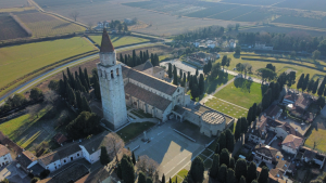 Zračni posnetek Bazilike v Ogleju (Basilica di Aquileia) z značilnim zvonikom, obkrožene z drevoredi in okoliško ravninsko pokrajino.