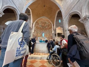 Un gruppo di visitatori all’interno di una chiesa si trova davanti all’altare decorato con ricchi affreschi; tra loro c’è anche una persona in sedia a rotelle e lo spazio presenta arcate in pietra.