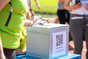 Oseba vlaga listek v škatlo za donacije; na škatli je QR koda za SMS donacijo.