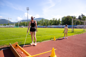 Odrasla oseba in otrok z zavezanimi očmi hodita po atletski stezi z belo palico in premagujeta ovire, označene s stožci.