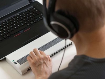 Oseba uporablja brajevo vrstico, povezano z računalnikom, za branje in navigacijo po digitalnih vsebinah.