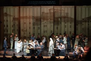 Spettacolo teatrale con numerosi attori sul palco e sopratitoli proiettati sopra la scena.